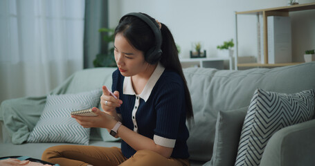 Selective focus of Asian teenager woman wear wireless headphones sitting on sofa making checklist of things to pack for travel, Preparation travel suitcase at home. - 772699551