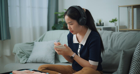 Selective focus of Asian teenager woman wear wireless headphones sitting on sofa making checklist of things to pack for travel, Preparation travel suitcase at home. - 772699568