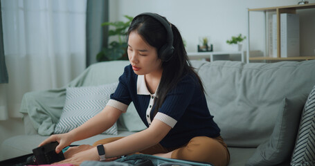 Selective focus of Asian teenager woman wear wireless headphones sitting on sofa packing travel luggage with personal items for traveling trip, Preparation travel suitcase at home. - 772699576