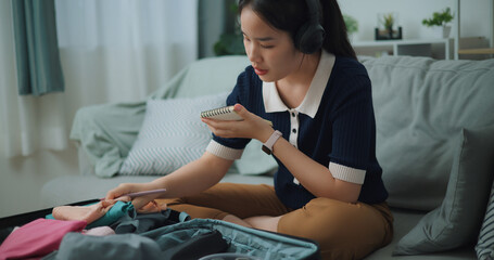 Selective focus of Asian teenager woman wear wireless headphones sitting on sofa making checklist of things to pack for travel, Preparation travel suitcase at home. - 772699593