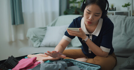 Selective focus of Asian teenager woman wear wireless headphones sitting on sofa making checklist of things to pack for travel, Preparation travel suitcase at home. - 772699705