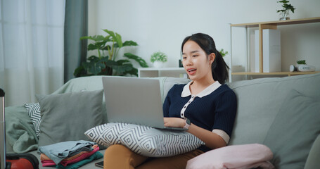 Portrait of Asian teenager woman sitting on sofa using laptop for prepare booking hotel and airplane ticket for travel. backpacker travel concept. - 772699716