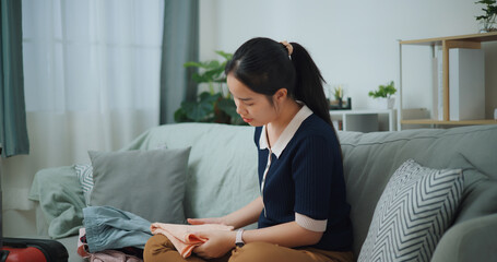 Side view of Asian teenager woman sitting on sofa packing travel luggage with clothes for traveling trip, Preparation travel suitcase at home. - 772699773