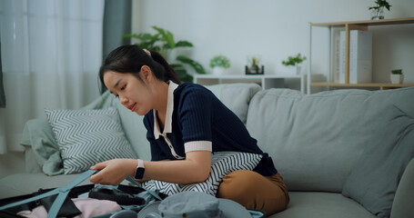 Side view of Asian teenager woman sitting on sofa packing travel luggage with clothes for traveling trip, Preparation travel suitcase at home.