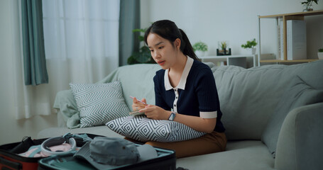 Side view of Asian teenager woman sitting on sofa making checklist of things to pack for travel, Preparation travel suitcase at home. - 772699924