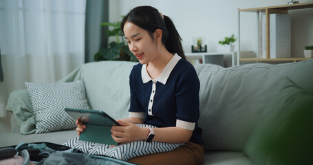 Portrait of Asian teenager woman sitting on sofa planning vacation trip and searching information on digital tablet, travel and lifestyle.