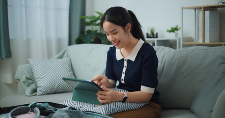 Portrait of Asian teenager woman sitting on sofa planning vacation trip and searching information on digital tablet, travel and lifestyle. - 772699970