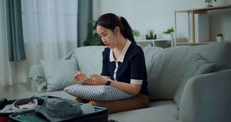 Side view of Asian teenager woman sitting on sofa making checklist of things to pack for travel, Preparation travel suitcase at home. - 772699974
