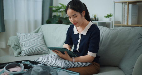 Portrait of Asian teenager woman sitting on sofa planning vacation trip and searching information on digital tablet, travel and lifestyle.