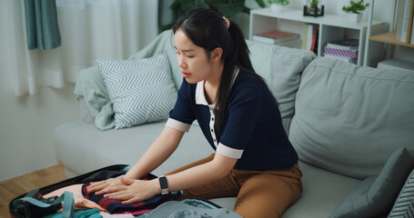 High angle view of Asian teenager woman sitting on sofa packing travel luggage with personal items...