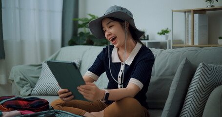 Portrait of Asian teenager woman sitting on sofa video call with friend while packing suitcase luggage for travelling, backpacker travel concept. - 772700337