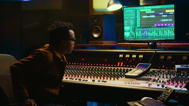 African american audio engineer uses compressor and mixer in professional studio, changing volume level and sound settings on a recording. Music production in control room. Camera A.
