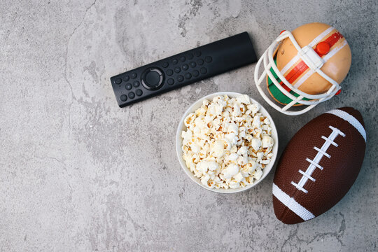 Top view of popcorn, rugby ball, helmet and remote control over grey background with copy space. Superbowl concept.