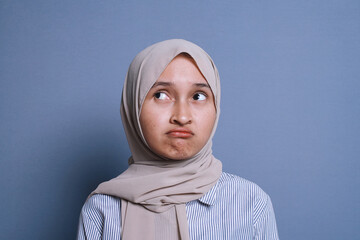 An upset muslim woman in hijab thinking about something and looking up while standing in the blue...