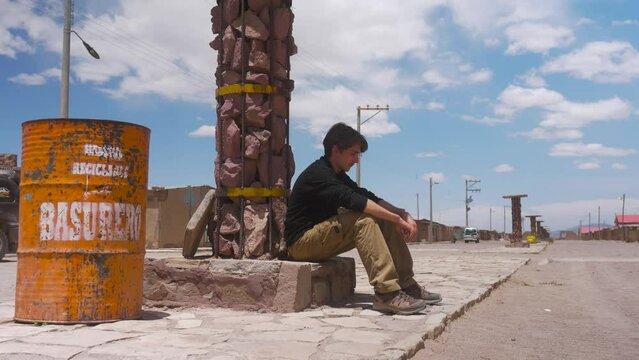 Lonely Man Sitting By Rock Column Next To Garbage Drum On Empty Street In Bolivia. wide shot