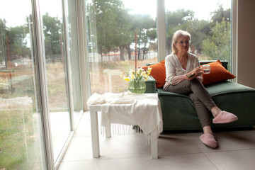 A beautiful elderly woman in a home outfit rests on the couch and drinks her hor drink
