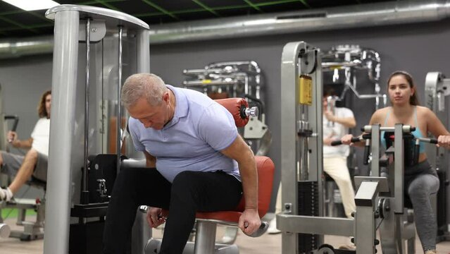 Elderly man training trains muscles of arms on the simulator in gym. High quality 4k footage