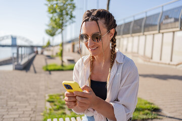 Positive hipster girl holds mobile phone, looks at cellphone screen, uses smartphone apps. girl with mobile phone on the street. 