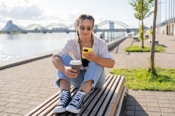hipster female with braids and takeaway coffee sitting using smartphone at street of city. Happy student girl sitting outside with smartphone in hands having coffee break have video call with friends.