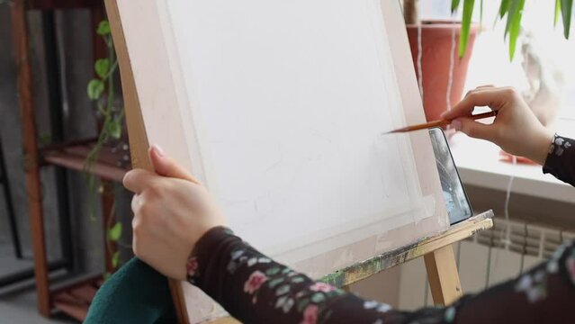 Female artist starting drawing with a pencil on paper in light art studio. Beautiful young woman paints on wooden easel. Blonde girl in black floral dress in creative process