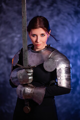 Fototapeta na wymiar Portrait of a young woman with scars on her face, dressed in black clothes with steel knightly hands and a gorget, holding a bastard sword, posing against an abstract background. Medieval fantasy girl