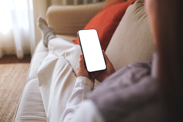 Mockup image of a woman holding mobile phone with blank desktop white screen while sitting on a sofa at home - obrazy, fototapety, plakaty