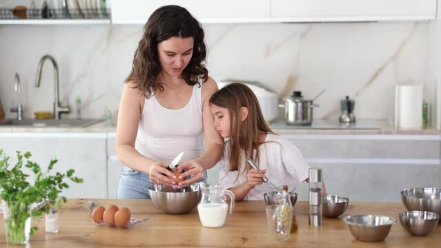 Mom teaches her daughter how to make dough, breaks eggs into bowl. Woman explains to her daughter successful recipe and rules for making dough for dessert. Homemade food and little helper.
