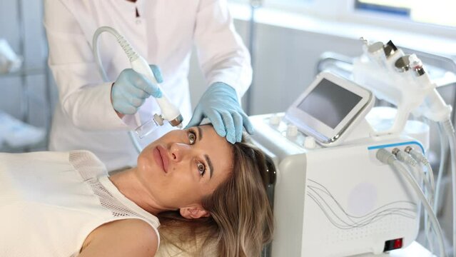 Concentrated young female patient undergoing facial procedure with application of atomized nutrients to enhance skin vitality at aesthetic medicine clinic. Modern technologies in hardware cosmetology