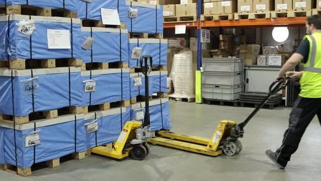 Warehouse worker in high-visibility vest walks between rows of blue-wrapped pallets, using a Hand Pallet Truck with Double Rollers