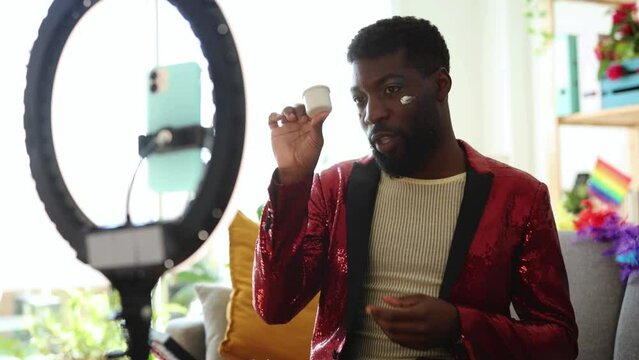 man doing tutorial for social media with smart phone for pride with makeup and ring light - A focused African American man in a glittering red sequin jacket applies facial cream, prepping for a Pride 