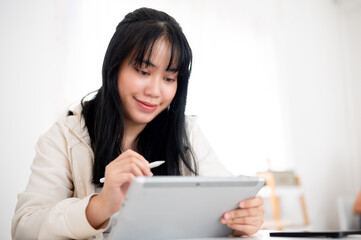 A young Asian woman using her digital tablet with a stylus pen at a table indoors.