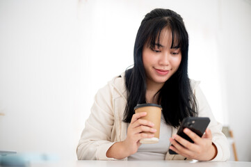 A positive, smiling Asian woman in casual wear is sipping coffee and using her smartphone in a cafe.