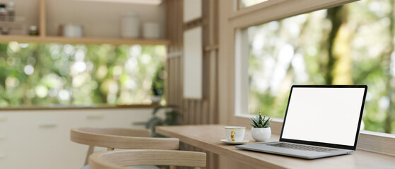A laptop computer mockup on a wooden dining table against the window in a neutral modern kitchen.
