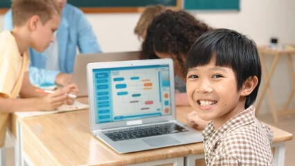 Energetic asian boy coding program and looking at camera in STEM class while caucasian teacher and...