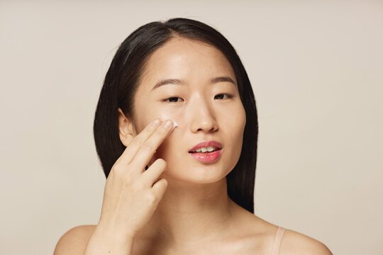 Studio portrait of young Asian woman applying nourishing or hydrating cream on her facial skin