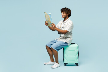 Full body traveler Indian man wear white casual clothes sit on bag read map isolated on plain blue...