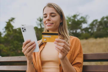 Fotobehang Close up young woman wears orange shirt casual clothes using mobile cell phone hold credit bank card sit on bench walk rest relax in spring green city park outdoors on nature. Urban lifestyle concept. © ViDi Studio