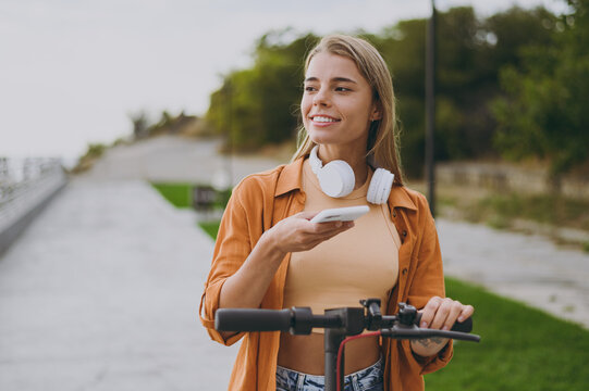 Fototapeta Young woman wear orange shirt casual clothes headphones riding e-scooter transport use mobile cell phone app walking rest relax in spring green city park outdoors on nature. Urban lifestyle concept.