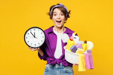 Young surprised happy shocked woman wear purple shirt hold in hand clock basin with detergent bottles do housework tidy up look camera isolated on plain yellow background studio. Housekeeping concept - obrazy, fototapety, plakaty