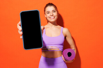 Young fitness trainer sporty woman sportsman wears purple top clothes in home gym hold yoga mat use blank screen mobile cell phone isolated on plain orange background. Workout sport fit abs concept.