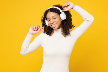 Little happy kid teen girl of African American ethnicity wear white casual clothes listen to music in headphones dance isolated on plain yellow background studio portrait. Childhood lifestyle concept.