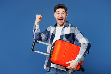 Traveler man wears shirt casual clothes hold suitcase bag do winner gesture isolated on plain blue background. Tourist travel abroad in free spare time rest getaway. Air flight trip journey concept.