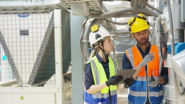 Professional Asian man and woman teamwork engineer in safety uniform working at outdoor construction site rooftop. Industrial technician worker maintenance checking building exterior air HVAC systems.