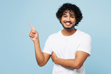 Young happy smiling Indian man he wear white t-shirt casual clothes point index finger aside on area look camera isolated on plain pastel light blue cyan background studio portrait. Lifestyle concept.