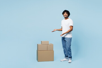 Full body side view young happy Indian man wear white t-shirt casual clothes point on stack cardboard blank boxes isolated on plain pastel light blue cyan background studio portrait Lifestyle concept - obrazy, fototapety, plakaty