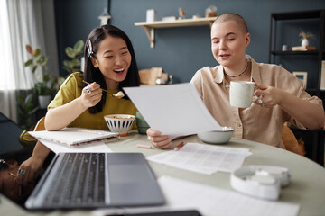 Two cheerful young Asian and Caucasian women sitting at table in kitchen eating breakfast and chatting about school project