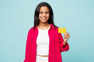 Little fun kid teen girl of African American ethnicity wear pink shirt white t-shirt hold mock up of credit bank card isolated on plain pastel light blue cyan background. Childhood lifestyle concept.