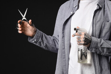 Young tattooed barber with hair spray and scissors on dark background, closeup