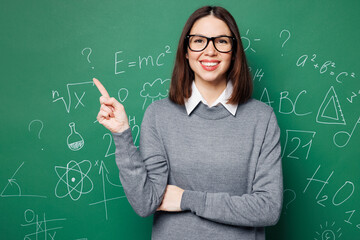 Young smiling smart teacher woman wear grey casual shirt glasses point index finger on blank area...