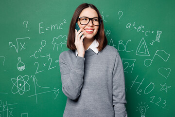 Young smiling smart teacher woman wears grey casual shirt glasses talk speak on mobile cell phone isolated on green wall chalk blackboard background studio. Education in high school college concept.